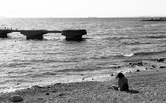 A lone woman sitting on a rocky beach, gazing at the sea in a black and white composition.
