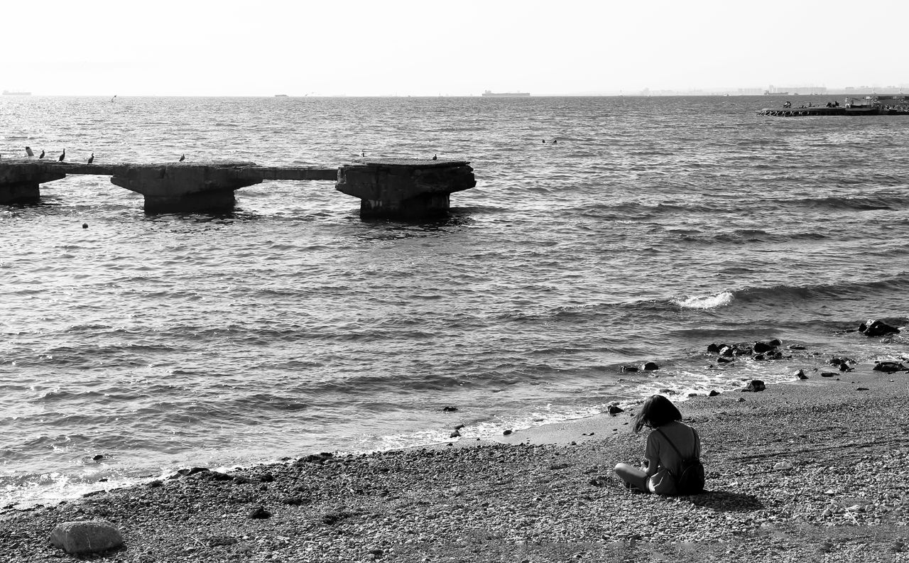 a lone woman sitting on a rocky beach gazing at the sea in a black and