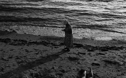 Free A contemplative senior woman in a dress and bucket hat stands on a sandy beach, using her smartphone, captured in a timeless black and white photograph. Stock Photo