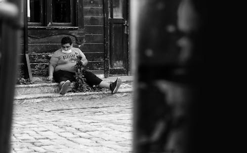 A Grayscale Photo of a Man Wearing Face Mask while Sitting on the Street