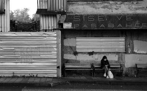 Grayscale Photo of Woman Sitting on the Wooden Bench near the Abandoned Building 