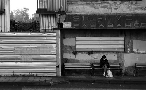 Free Black and white photo of a woman in a mask sitting on a street bench, urban decay setting. Stock Photo