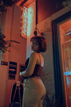 Young woman posing under neon lights outside a tattoo shop at night, showcasing urban nightlife vibes.