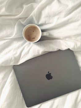 A cozy morning featuring a coffee cup and laptop on soft white bedding, viewed from above.