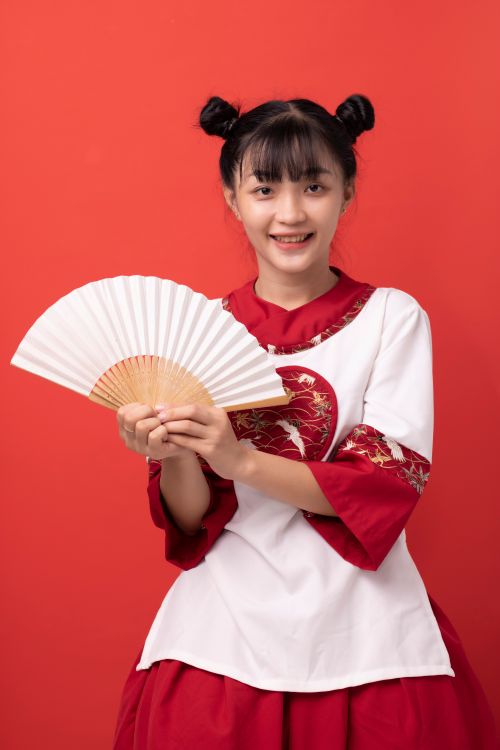 Free Asian teen girl smiling and holding a fan, wearing traditional attire against a red background. Stock Photo