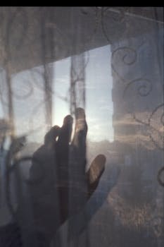 A silhouette of a hand pressed against a window with a dreamy dawn sky.