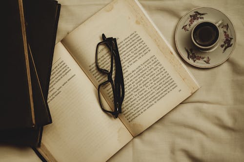 An Open Book with Eyeglasses Near the Cup of Coffee on Ceramic Saucer