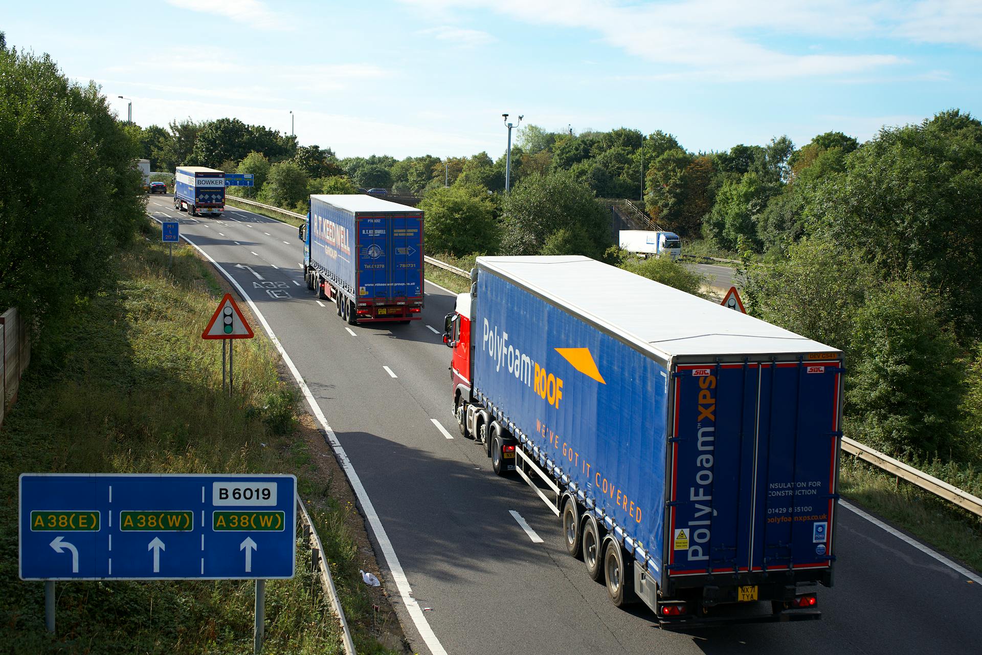 Cargo trucks on A38 highway, Mansfield, UK, surrounded by summer greenery and clear skies.
