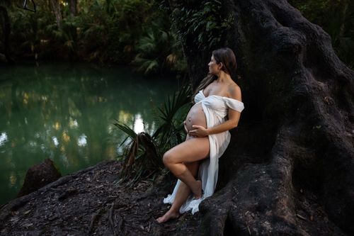 Free A pregnant woman in a white dress poses by a tree trunk in a tranquil forest setting. Stock Photo