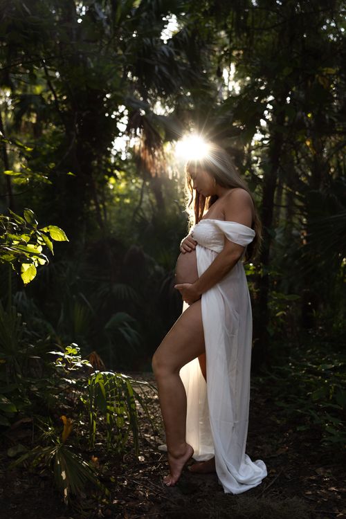 Free Pregnant woman in white dress poses amidst sunlit forest setting. Stock Photo