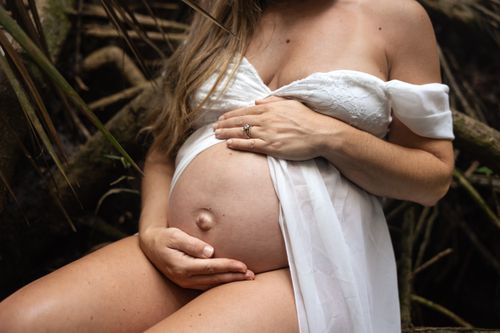 Free A serene outdoor maternity photoshoot capturing a woman in a white dress cradling her baby bump. Stock Photo