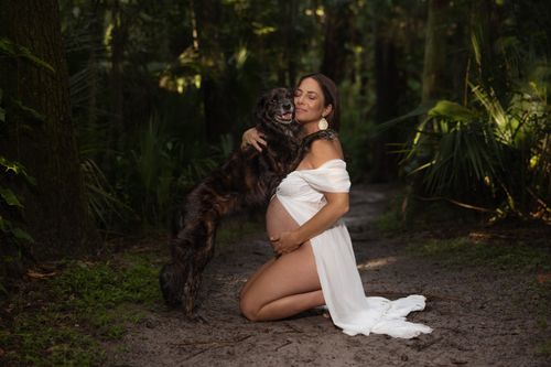 Free Pregnant woman in white dress hugging dog in a serene forest environment. Stock Photo