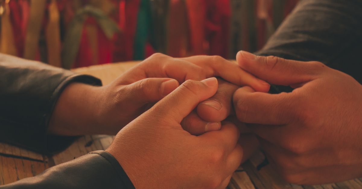 Warm and intimate close-up of a couple holding hands on a wooden table with colorful ribbons in background.
