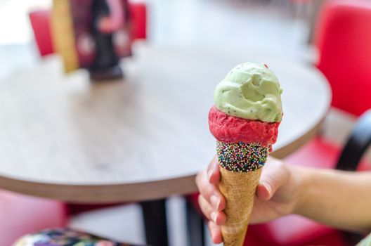 Hand holding a colorful ice cream cone topped with mint and raspberry scoops indoors.