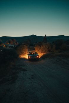 A car drives through a dark countryside path with headlights illuminating the way beneath a serene sky.