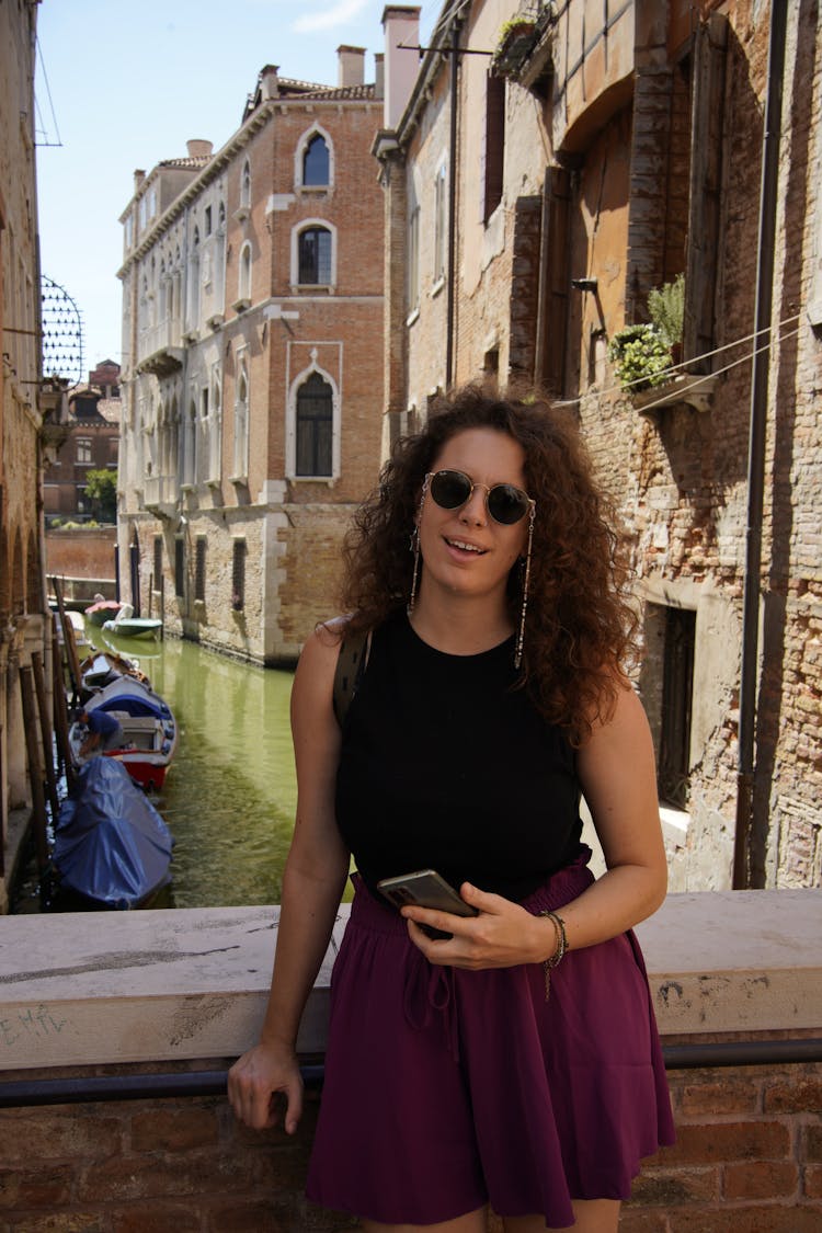 A Woman In Black Tank Top And Purple Skirt Wearing Sunglasses While Standing Near The Canal Between Buildings