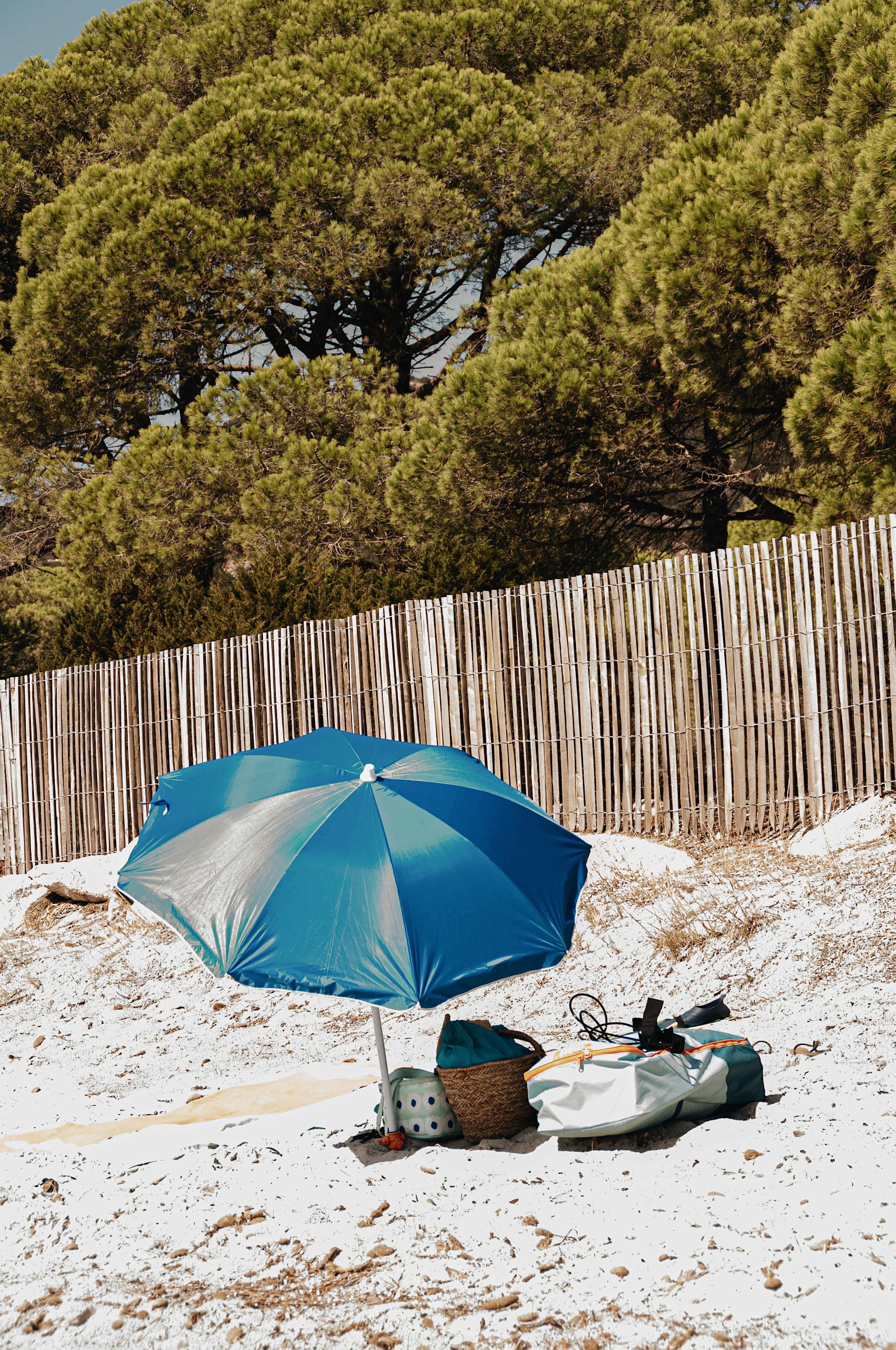 A tranquil beach setting in France with a blue umbrella, bags, and a wooden fence.