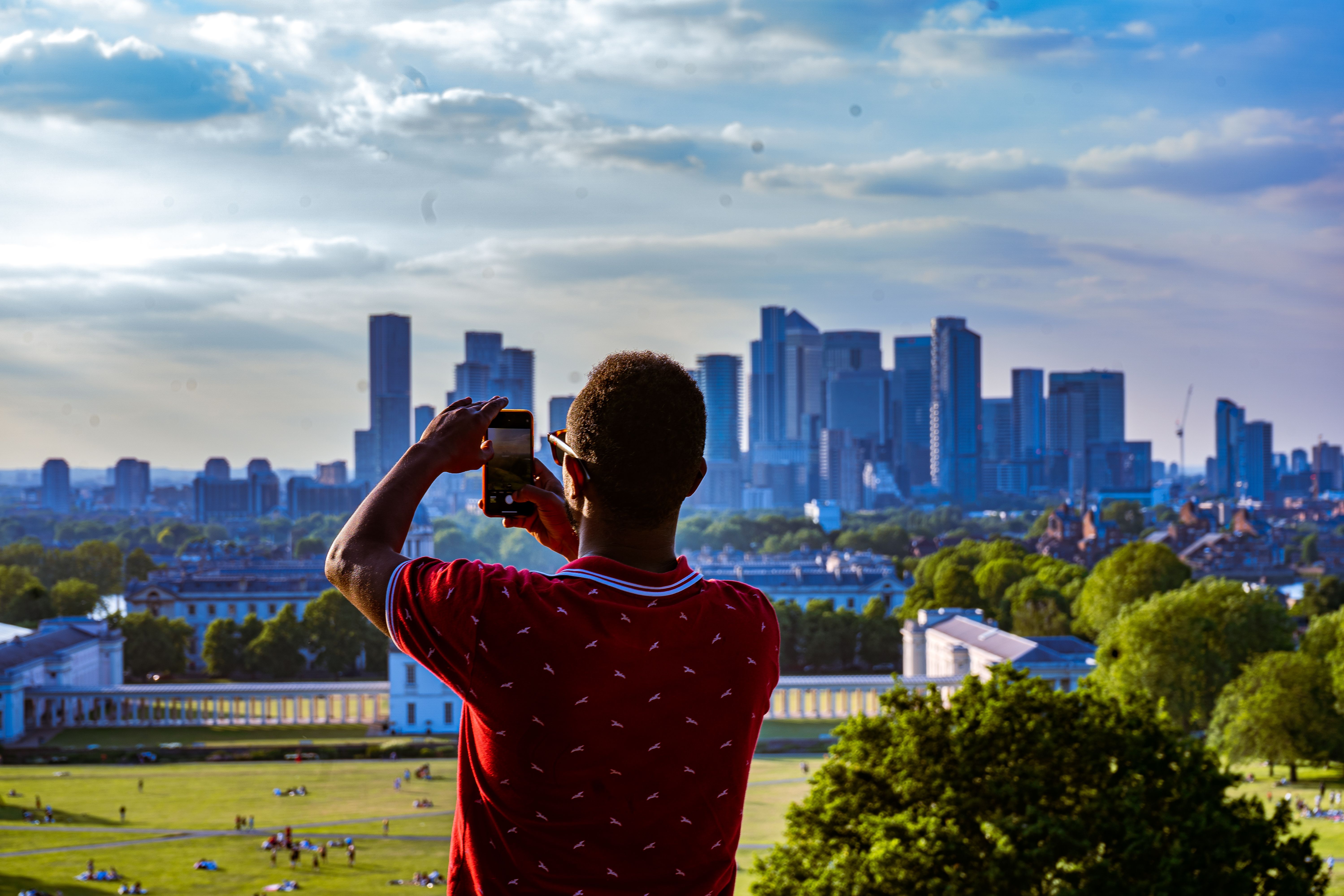 A man takes a picture of London's cityscape with his smartphone from a park on a sunny day