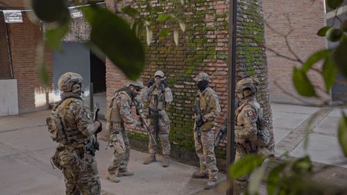 Free A team of soldiers in camouflage uniforms and gear stand strategizing by a mossy brick wall. Stock Photo