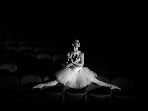 Grayscale Photo of Woman in Tutu Dress Posing on Top of Theatre Seats