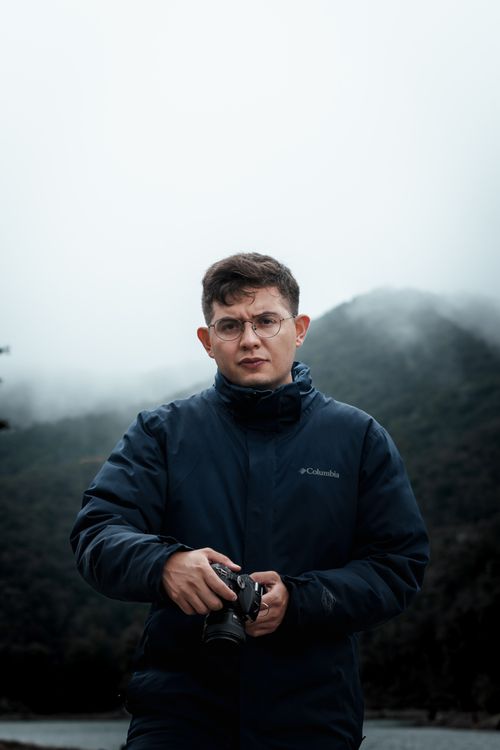 Free Portrait of a young man holding a camera, standing in a misty outdoor setting. Stock Photo