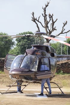 A Bell 407 helicopter with open doors, grounded outdoors with visible pilots and trees in the background.