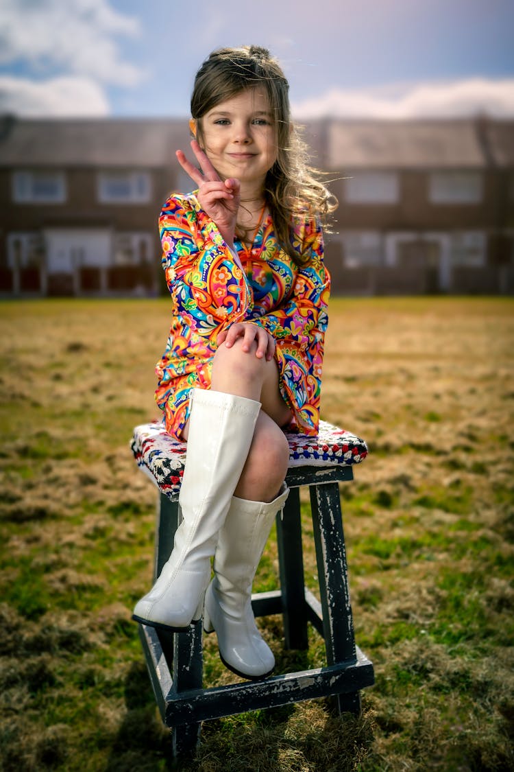 Cute Young Girl Sitting On A Wooden Stool And Doing A Peace Sign 