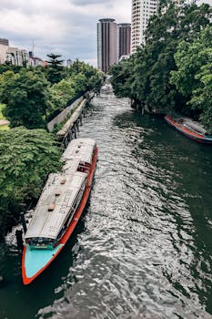 Explore urban Bangkok with this aerial view of a canal lined with boats and lush trees.