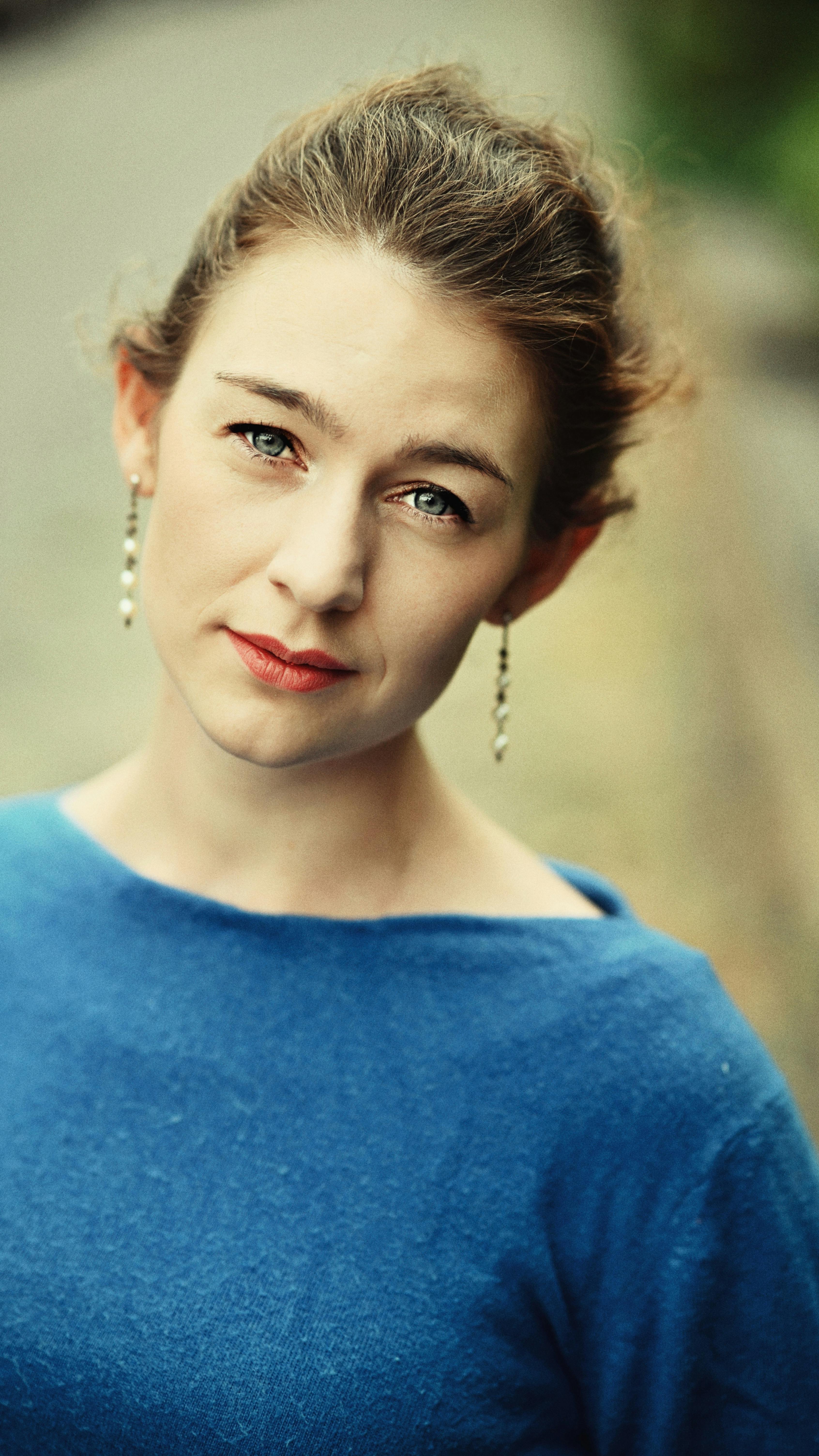 An elegant portrait of a woman with light eyes wearing a blue sweater and drop earrings outdoors.