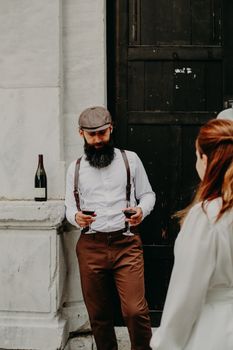 A bearded man in vintage attire holds wine glasses outdoors by a rustic door.