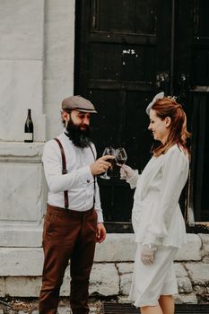 Stylish couple raising glasses of red wine in a celebratory outdoor setting, embracing vintage elegance.