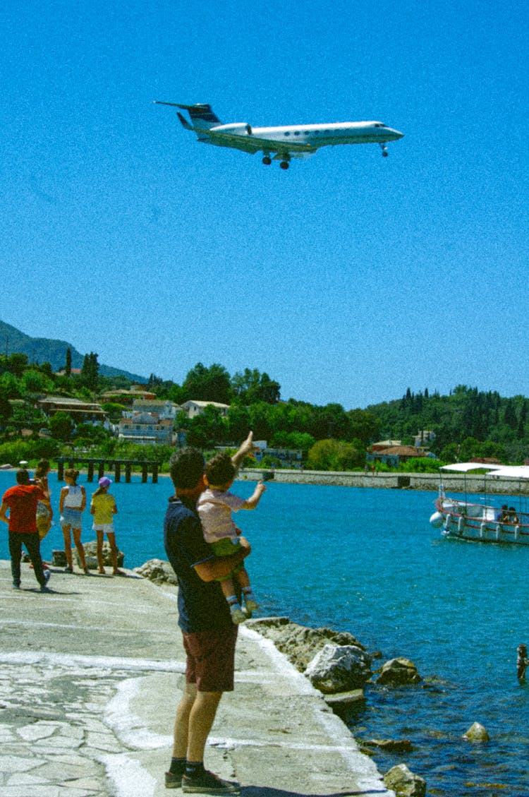 Father Holding A Child And Pointing At An Airplane Flying Above Them 