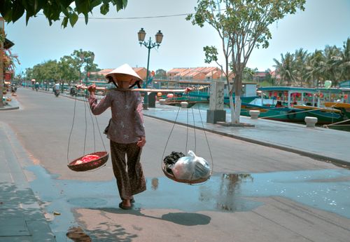 Free A woman wearing a Vietnamese hat carries groceries on a street, using a traditional carrying pole. Stock Photo