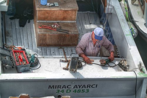 Free A man repairs equipment on a fishing boat at a dock, surrounded by tools and gear. Stock Photo