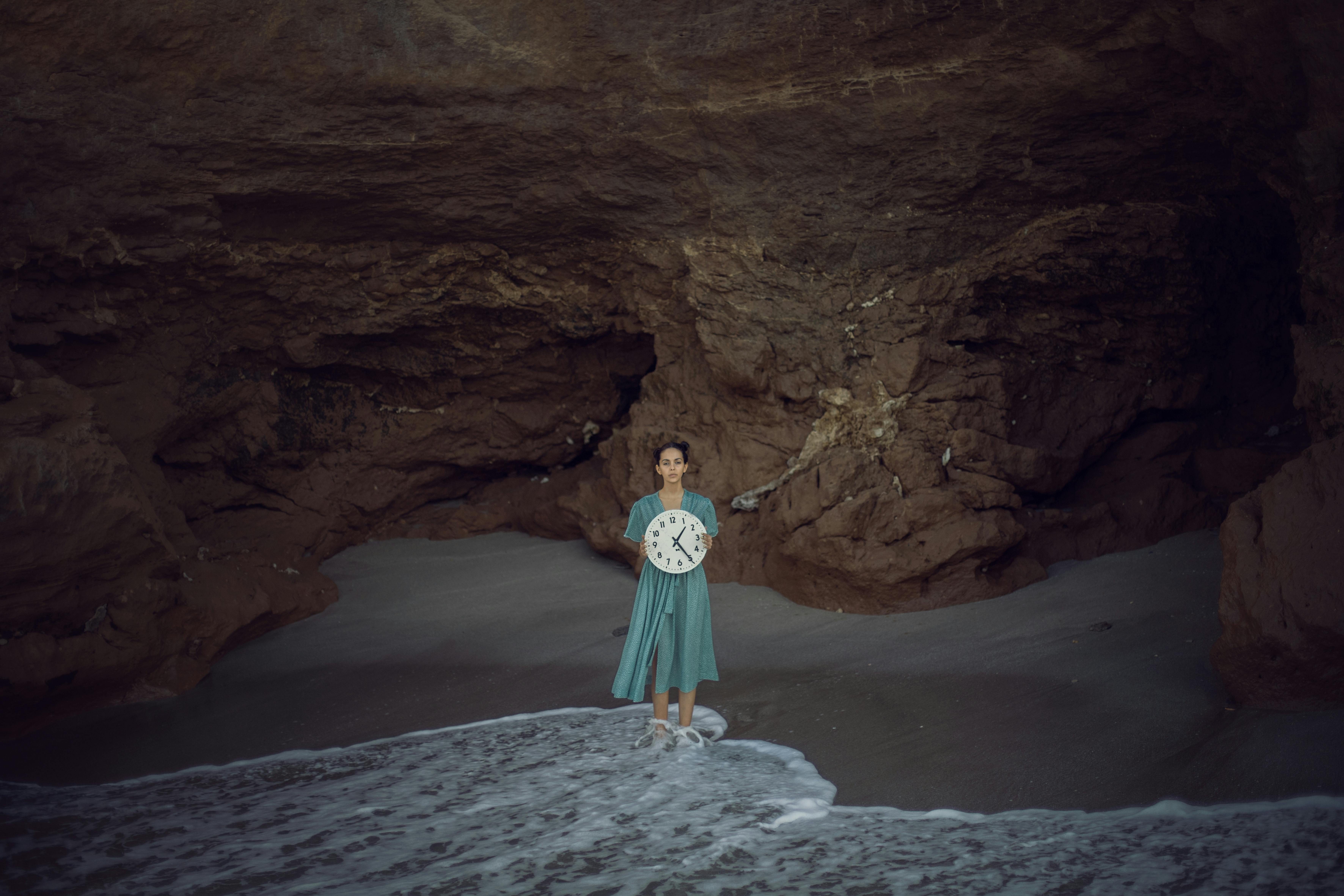 A woman stands with a clock in a tranquil cave by the sea, embodying timelessness and nature.