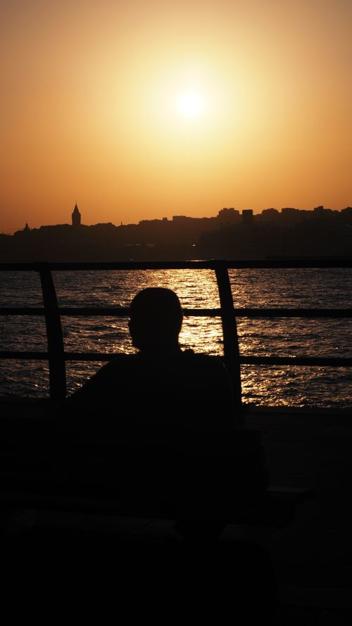 Free A silhouette of a person sitting by the sea during a stunning sunset with an orange sky. Stock Photo
