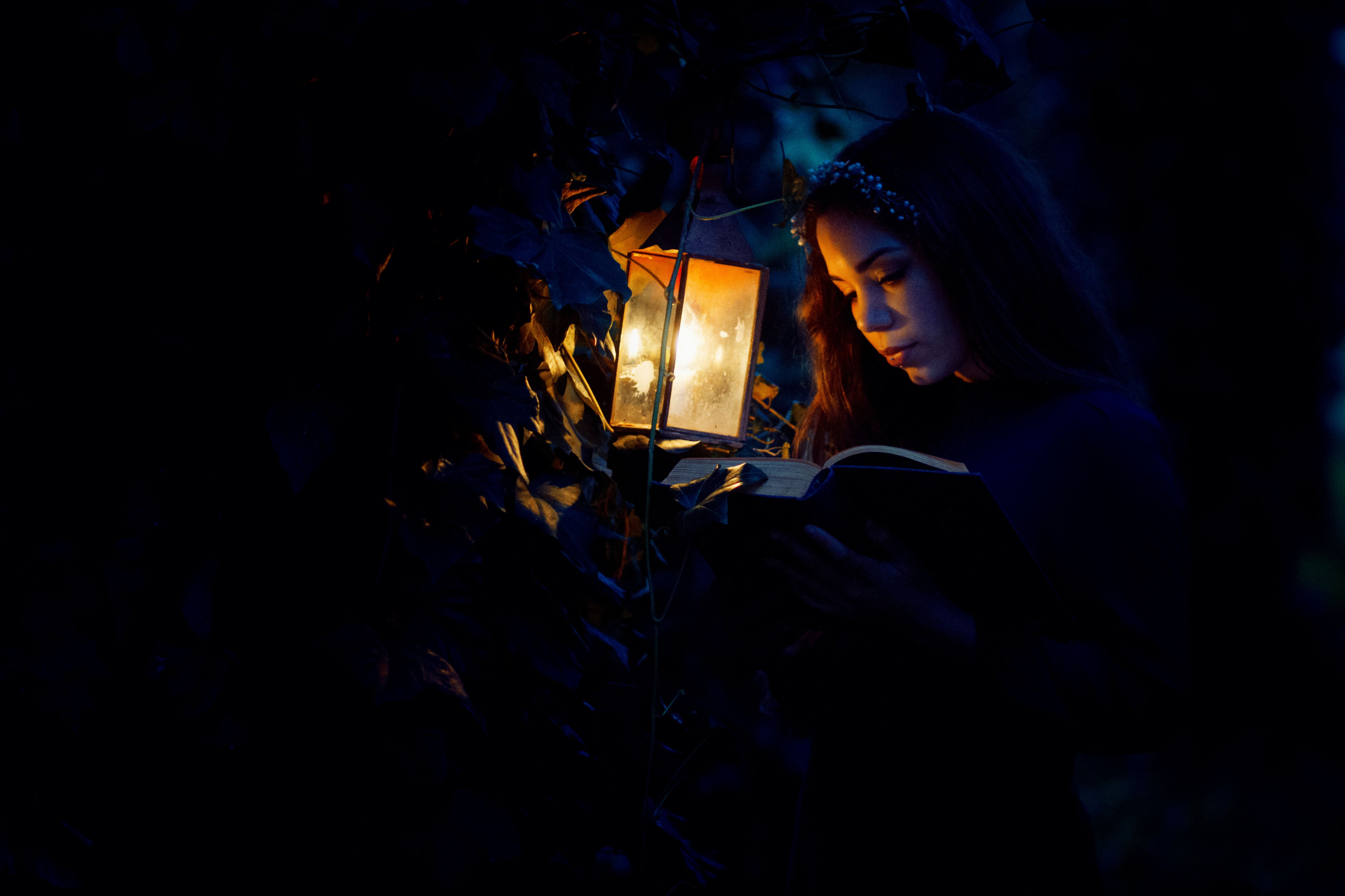 A woman deeply engrossed in a book, illuminated by a warm lantern in a dark forest setting.