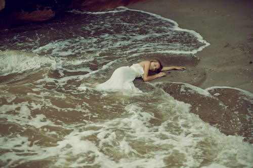Woman in a White Dress Lying on a Seashore with Waves Splashing on Her 