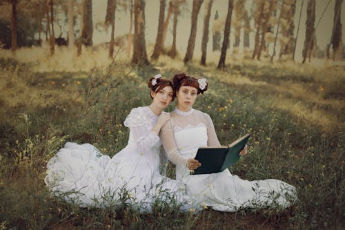 Two Women in Long White Old-fashioned Dresses Sitting on the Ground in the Forest and Holding a Book 
