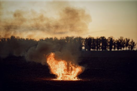 A solitary bonfire burns intensely on a vast open field, silhouetted by the evening sky.