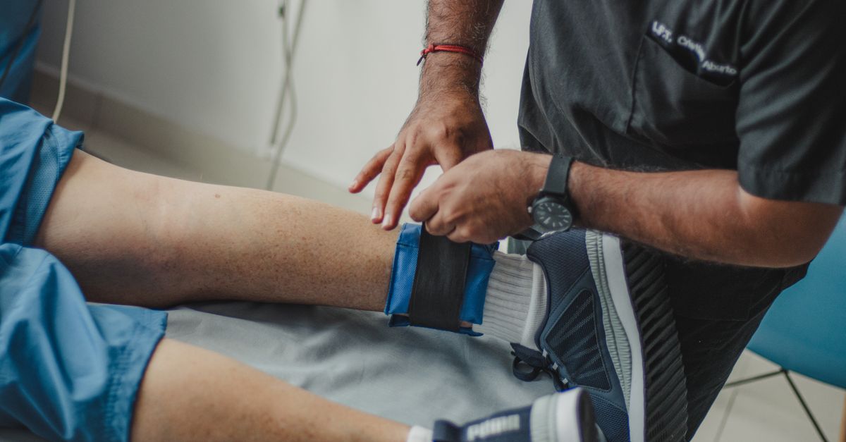 A physiotherapist adjusts a leg strap on a patient in a clinical setting.