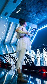 Model dressed in white cosplay attire holding a lightsaber indoors, surrounded by futuristic decor.