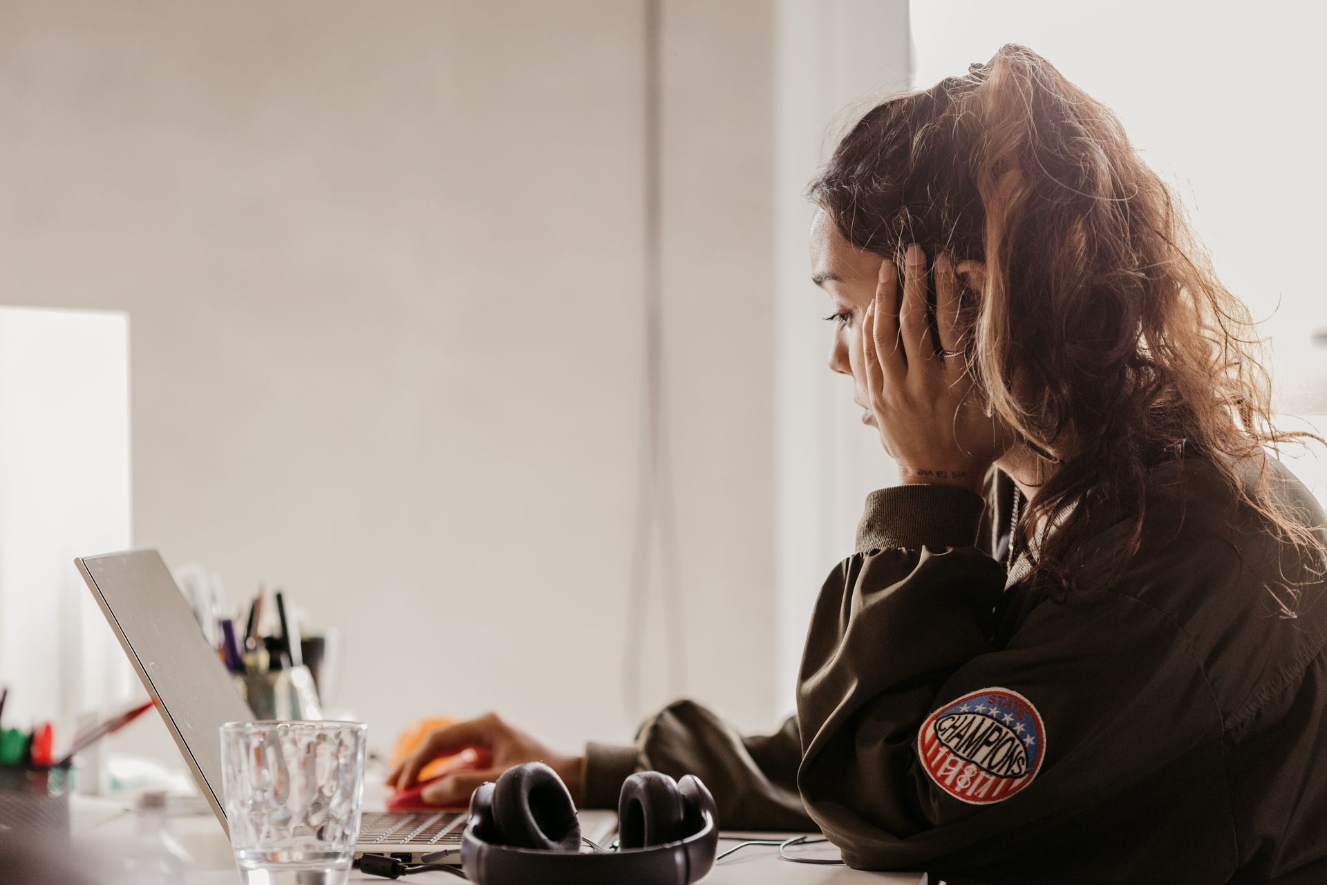 Young woman in a casual jacket using a laptop at a desk with headphones nearby.