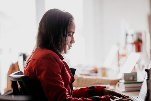 Focused young woman in red jacket typing on laptop in bright workspace.