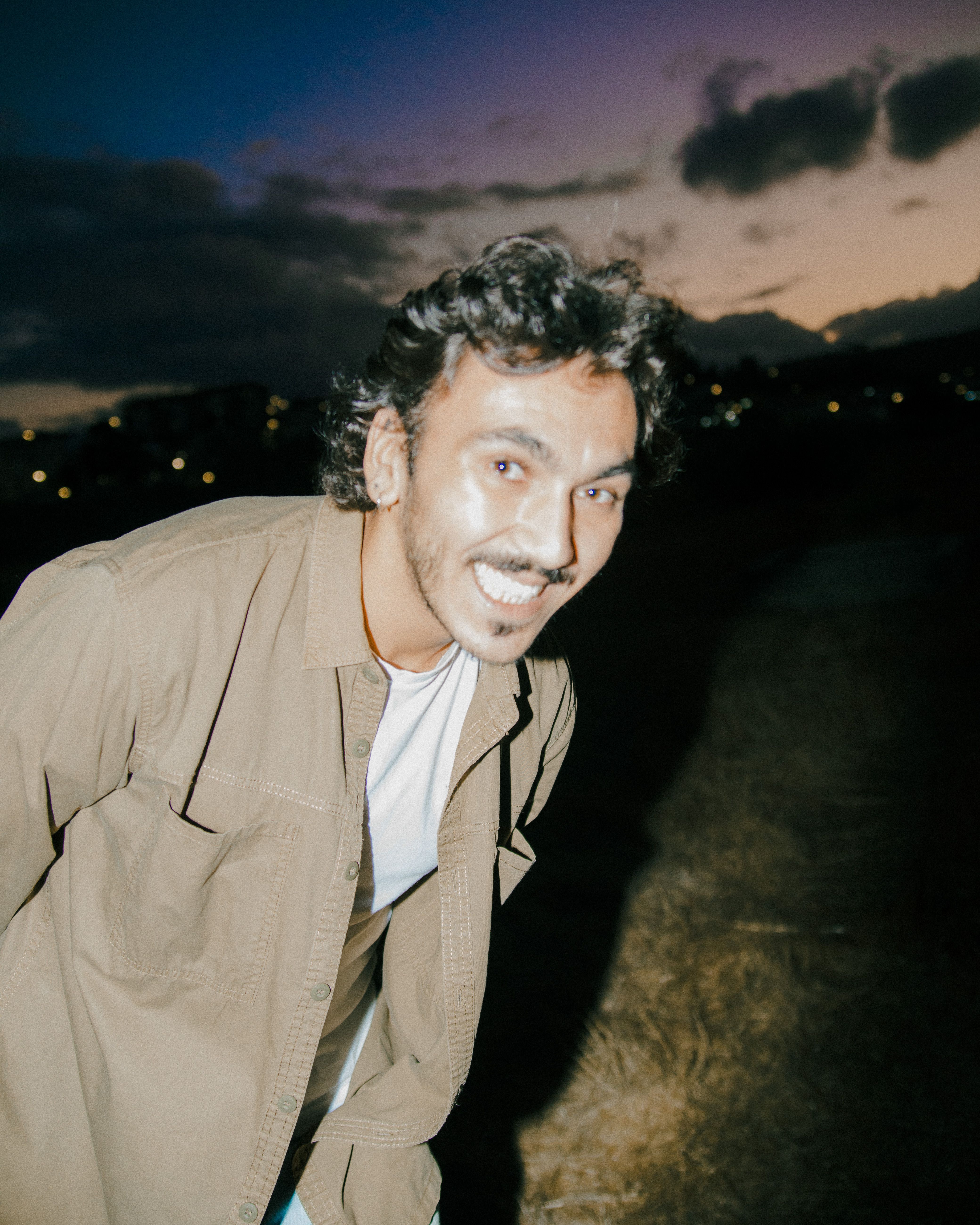 A cheerful young man with a mustache smiling outdoors at dusk.