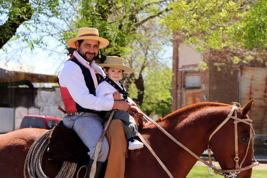 A father and son in traditional attire enjoying a horseback ride in a sunny outdoor setting.