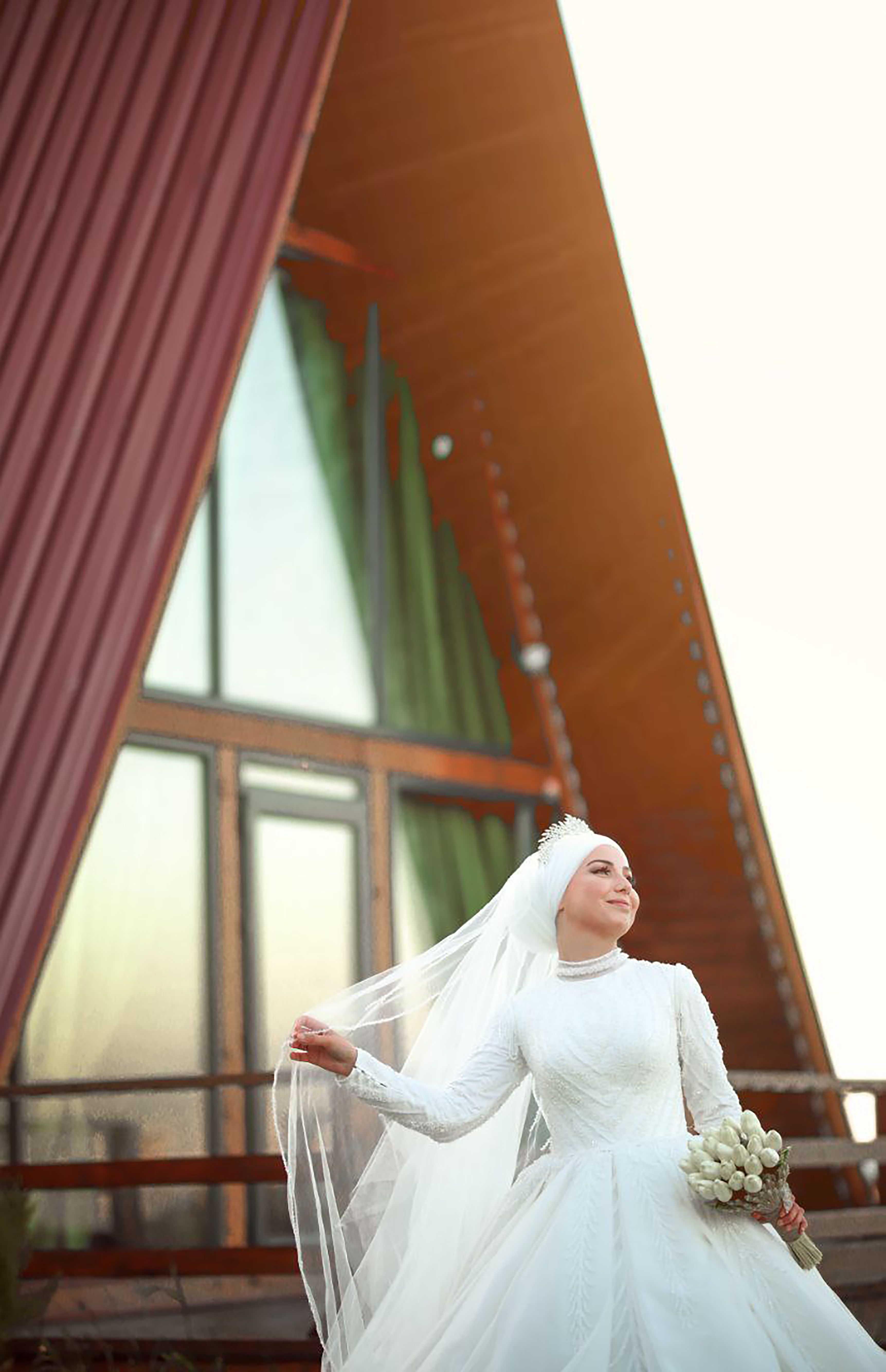 A smiling bride in a white dress and veil stands outdoors, holding flowers near a modern A-frame house.