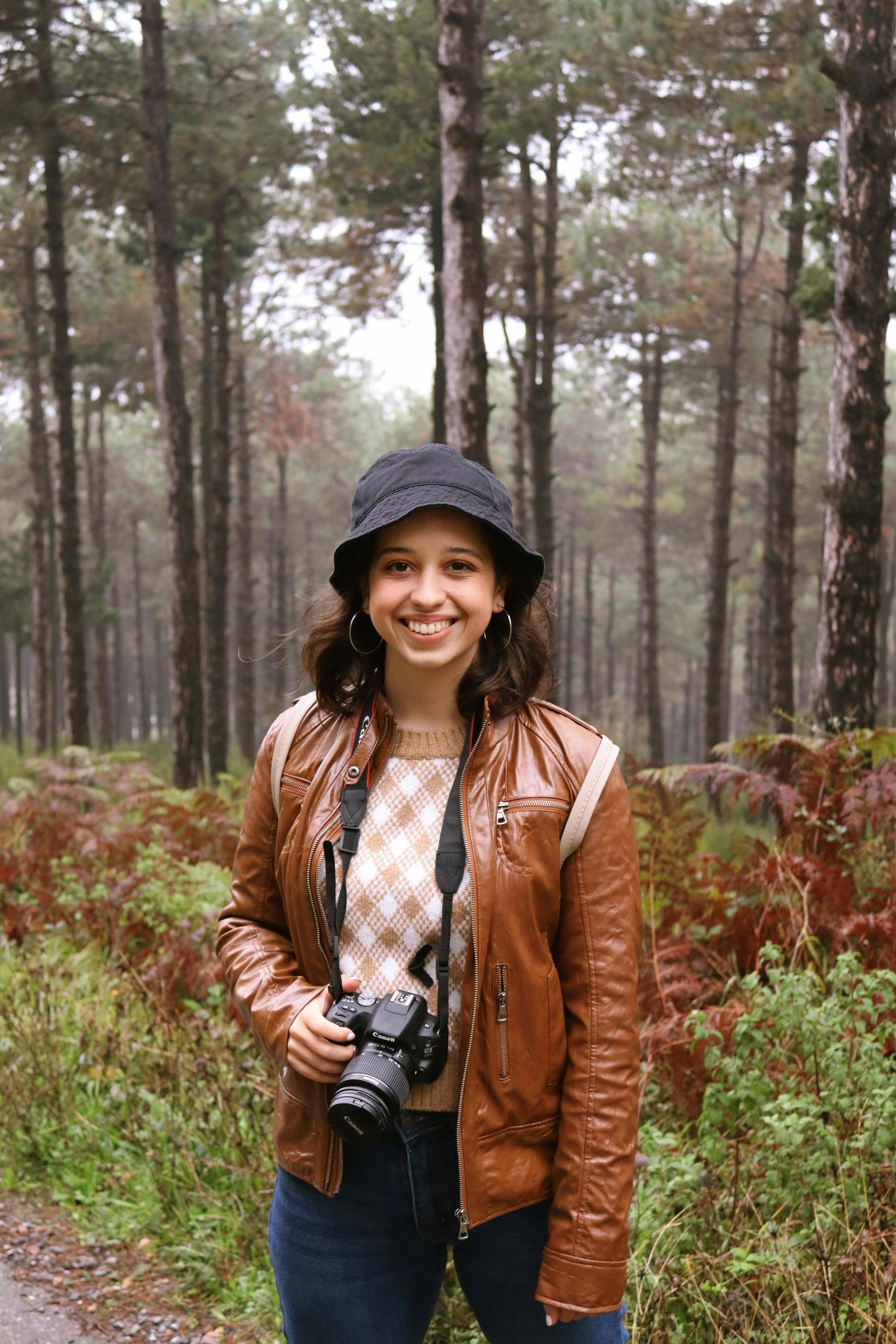 Smiling woman holding a camera in a misty forest, wearing a brown leather jacket and bucket hat.
