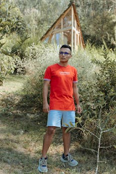 Man in casual attire poses in a lush garden setting with a rustic hut in Cáqueza, Colombia.