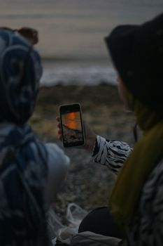 Two women view a sunset on a smartphone while sitting on a rocky beach.